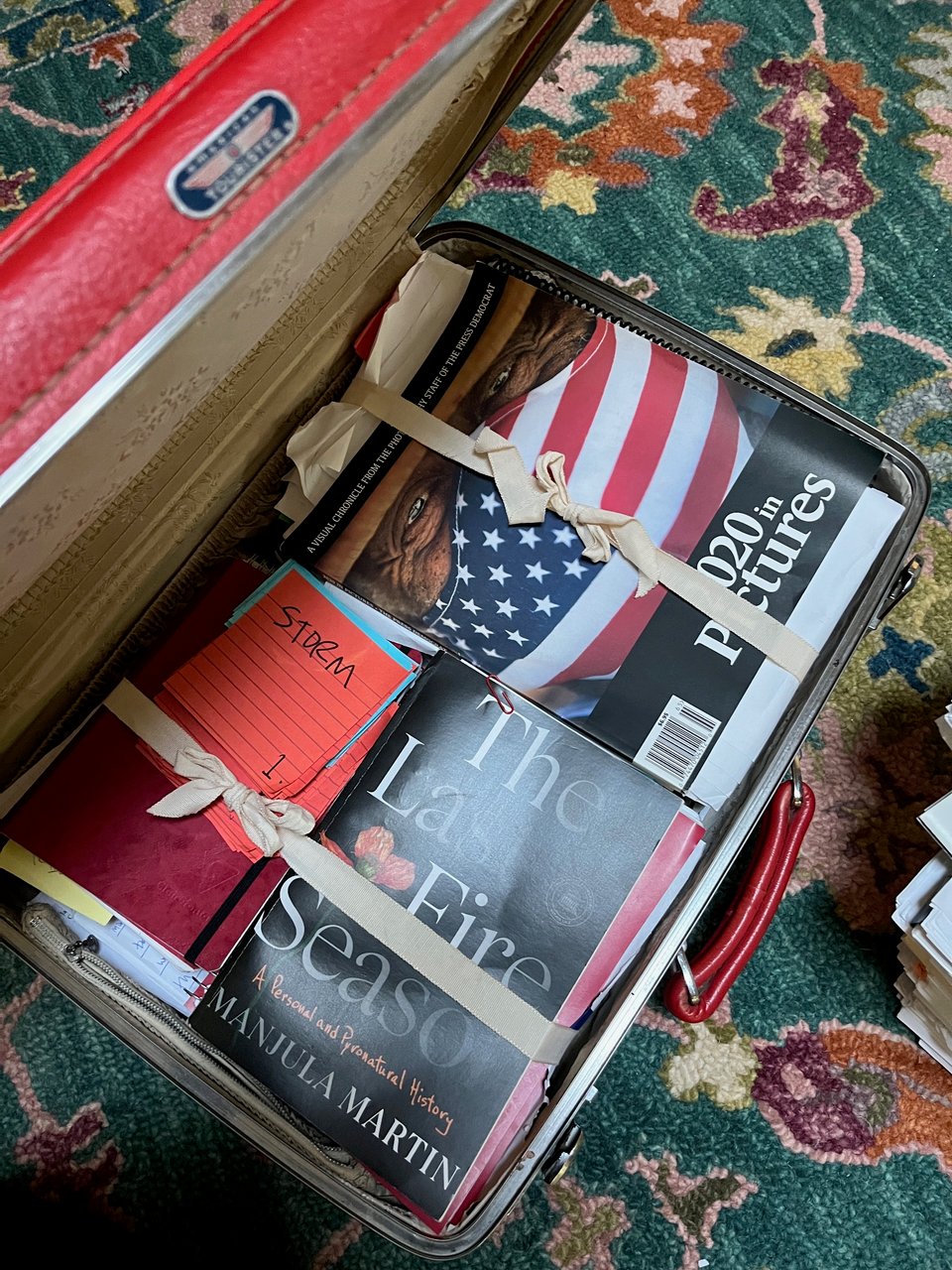 picture of a vintage suitcase full of papers and old manuscript drafts, on top a galley of The Last Fire Season and a magazine image of a white man in an American flag facemask, above him the text "the year in pictures, 2020"