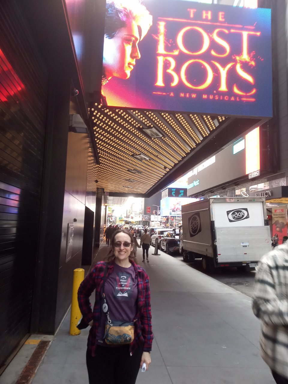 The author, wearing a T-Shirt with the cover of her novella THE NIGHT LIBRARY OF STERNENDACH on it, by the Palace theater in NYC, under an electronic poster for THE LOST BOYS.
