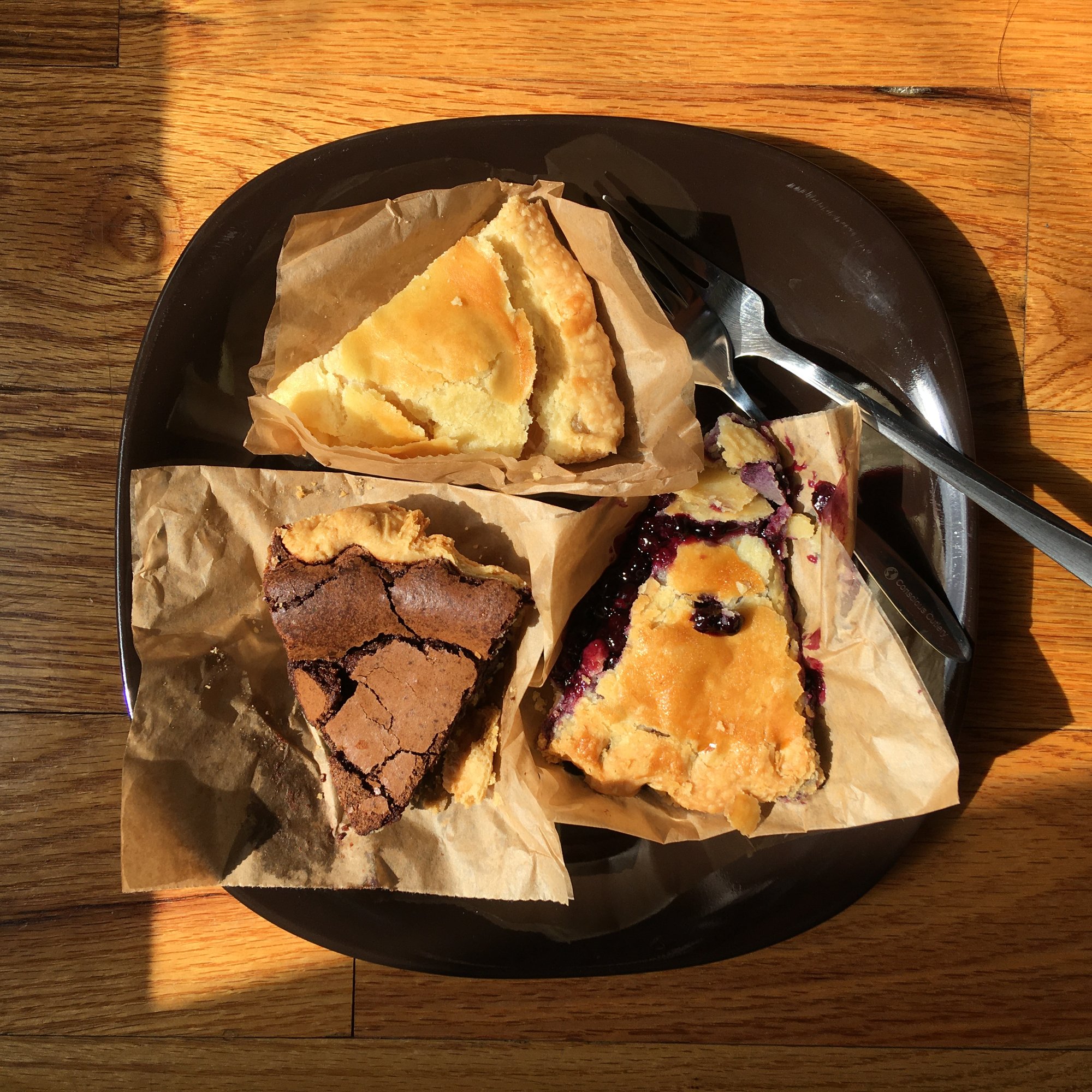 Photo depicts an overhead shot of three slices of pie: chocolate, apple, and blueberry. The pies sit on a dark brown plate, which rests on a wooden floor in a slice of afternoon light.