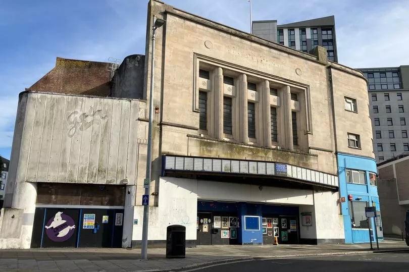 A 1930s moderne cinema with 1960s additions and later changes. The central building is dressed in portland stone and has large columns and long windows. To the right, part of the building is painted sky blue. To the left, a 1960s Gala bingo hall entrance has been added. The sign itself has gone but the weathering of the concrete still shows it.