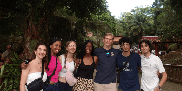 A group of seven young adults poses together in a lush, green park with trees and palm fronds surrounding them. They are standing close, smiling, and appear to be enjoying their time outdoors.