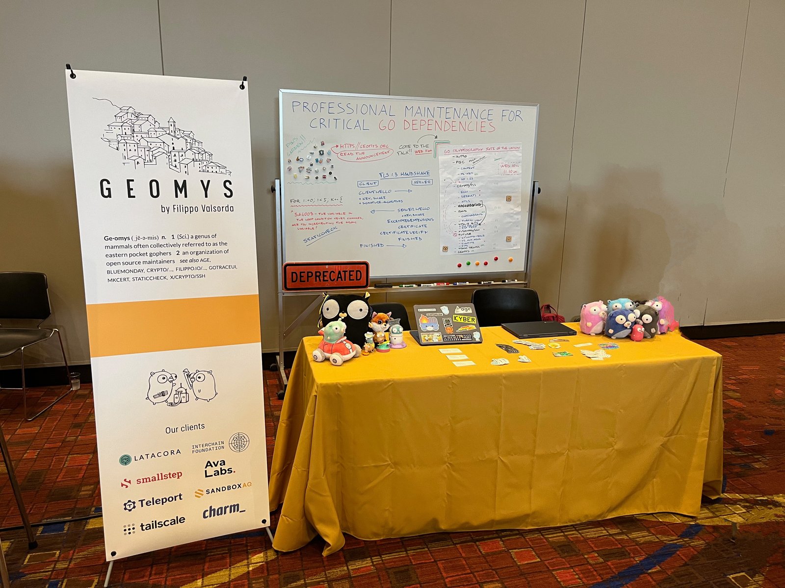 A photo of a conference booth. On the left, there's a tall white and yellow vertical banner. The top half has the Geomys logo. Below that is a description of Geomys. The bottom half of the banner lists Geomys clients. In the center, there is a table covered with a yellow tablecloth. On the table, there are several plush gophers. Some are black, while others are various pastel colors like pink, blue, and purple. In the middle of the plush toys, there are some some stickers and a laptop. Behind the table, a large whiteboard with handwritten notes and diagrams, including the TLS 1.3 handshake, and a little garden of enamel pins. A metal street sign with the word "DEPRECATED" is leaning against the bottom of the whiteboard.