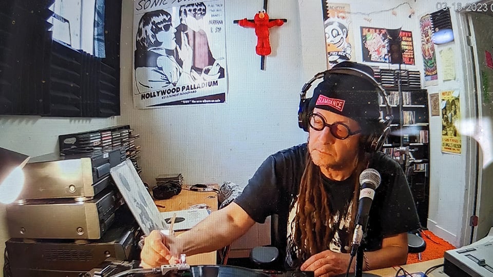 A man with dreadlocks and glasses and a beanie and headphones sits amidst much stereo gear as he sets the needle on a record.