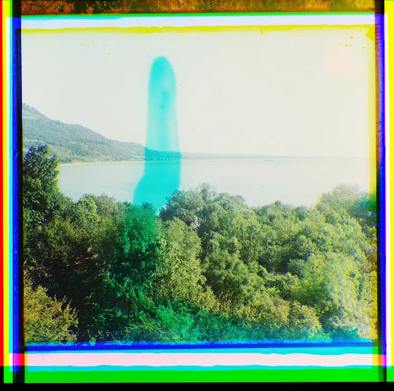 An experimental photograph showing a lush lakeside landscape overlaid with a ghostly, semi-transparent cyan form standing in the center. Colorful bands perhaps of film registration error frame the edges.