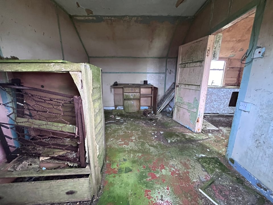 A derelict croft house interior with wooden board walls stained green with paint peeling from the walls. A desk without drawers stands in the corner with a closet bedframe near the camera. You can see the sink in the next room with some naked plywood.
