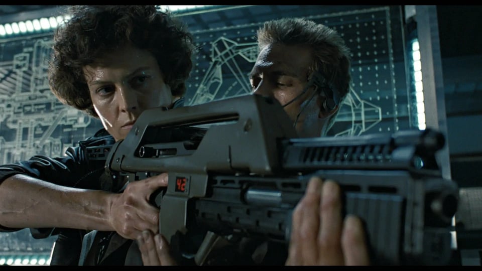 Ripley trains with a rifle and grenade launcher with Hicks looking on