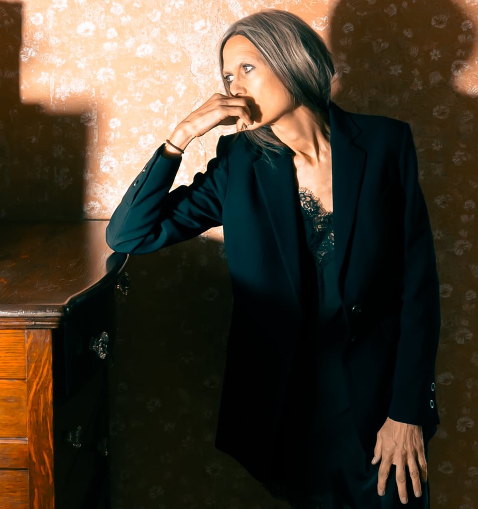 A woman in a black blazer with gray-blond hair looks to her right, with her right hand resting on her mouth and right elbow on a wooden table or dresser.