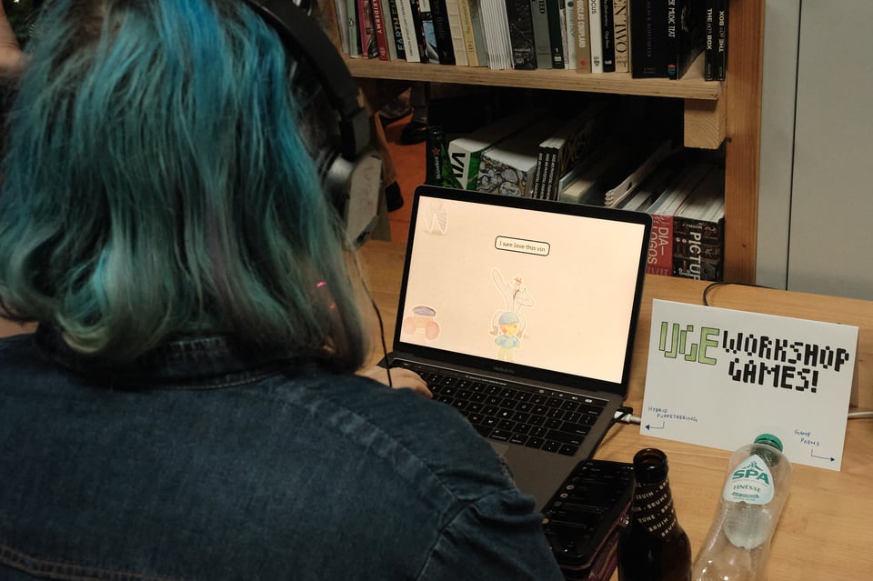 Person with blue hair plays a game next to a sign that says "UGE Workshop Games"