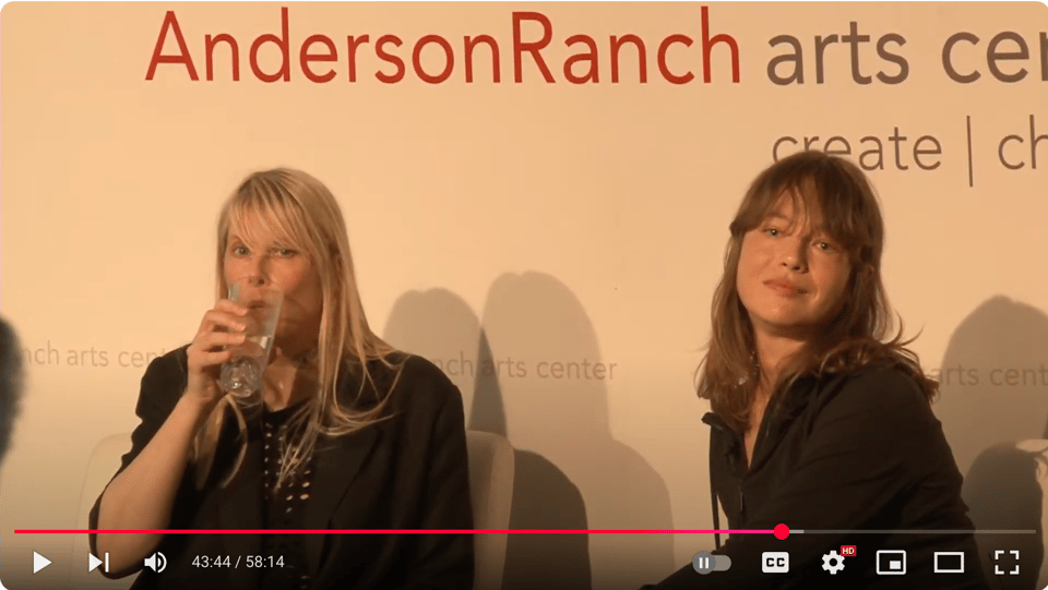 Johanna Fateman drinks water from a glass while Issy Wood gazes beatifically at the viewer. They are seated together, participating in a public conversation. Signage behind them reads "AndersonRanch."