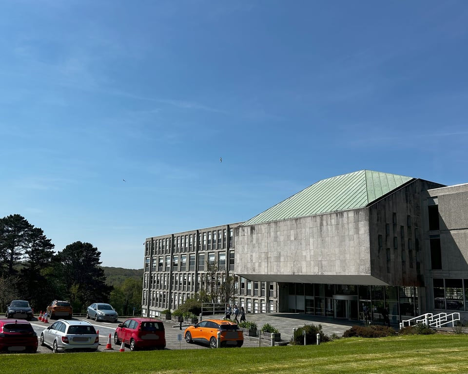 A large brutalist building with a carpark, overlooking a deep, wooded valley. The repetition of the grid of office windows is disrupted by the heavy block of a cantilevered chamber.