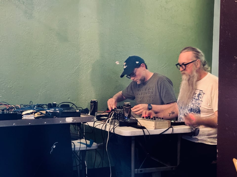 Mandom sitting side by side performing with their electronic instruments