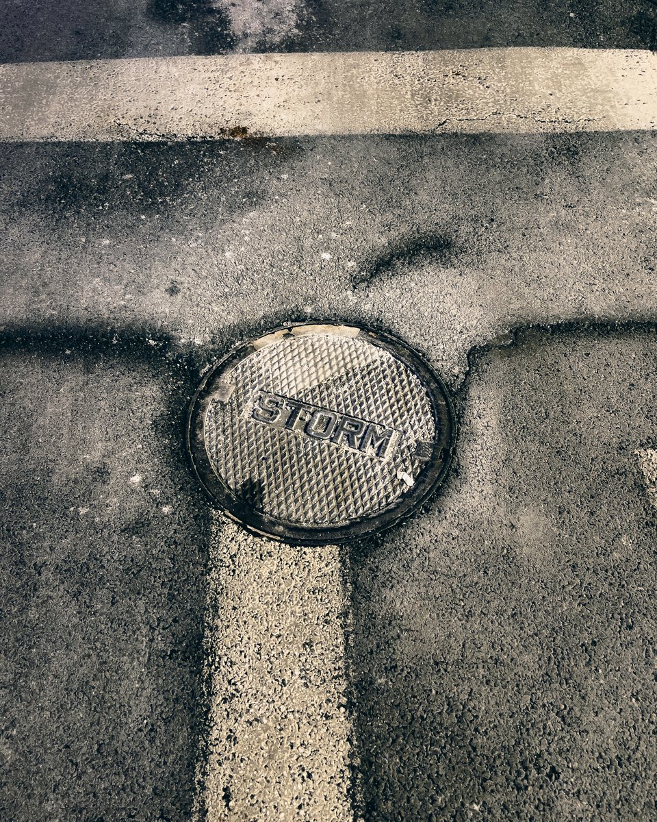 a manhole cover embossed with the word storm sits at the top of a broad vertical white stripe road marking, another white stripe crosses the frame horizontally at the top