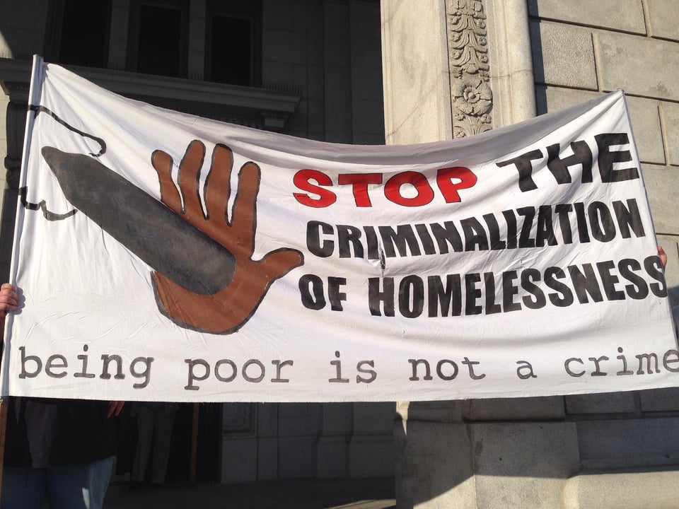 A banner in front of City Hall says STOP THE CRIMINALIZATION OF HOMELESSNESS: being poor is not a crime. There's a brown hand with stopping looks like a bullet
