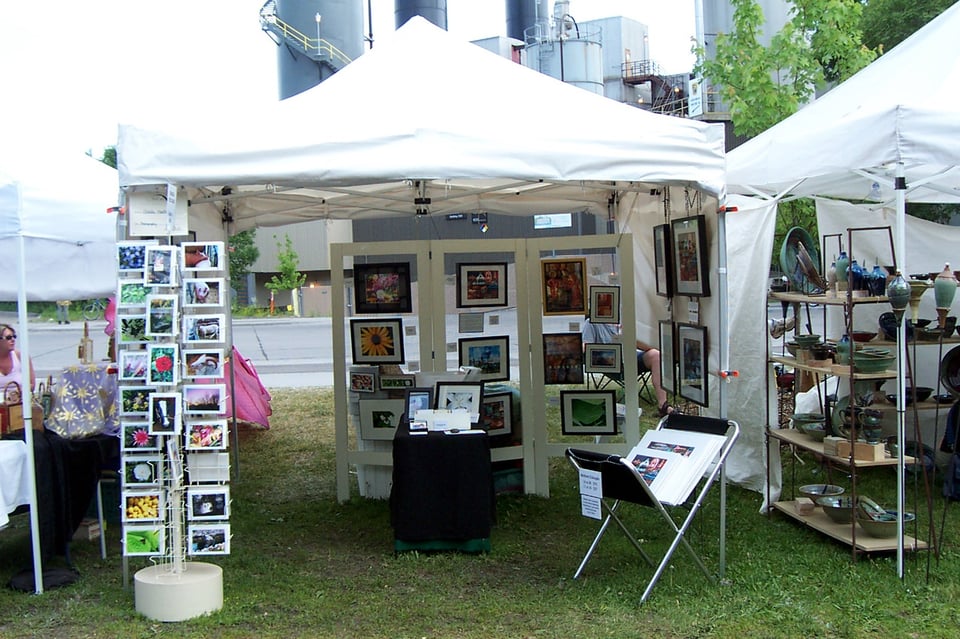 White tent at art fair on grass. Card rack and print rack sit outside,  folding display and table inside with framed art.