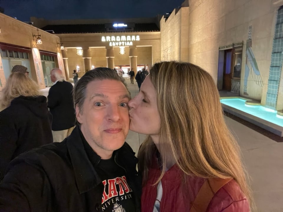 A man and a woman stand outside a theater. The sign in the background reads Grauman's Egyptian. A woman kisses a man on the cheek and he stares happily at the camera.