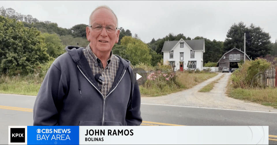A man stands in front of a farm house and a barn and reports the news.