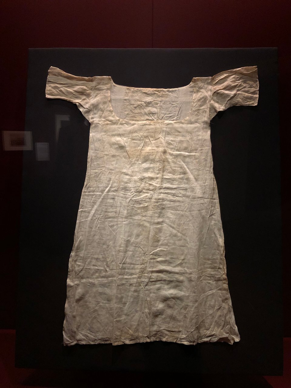 A very simple, slightly stained and crumpled, linen garment in a dark exhibition cabinet.