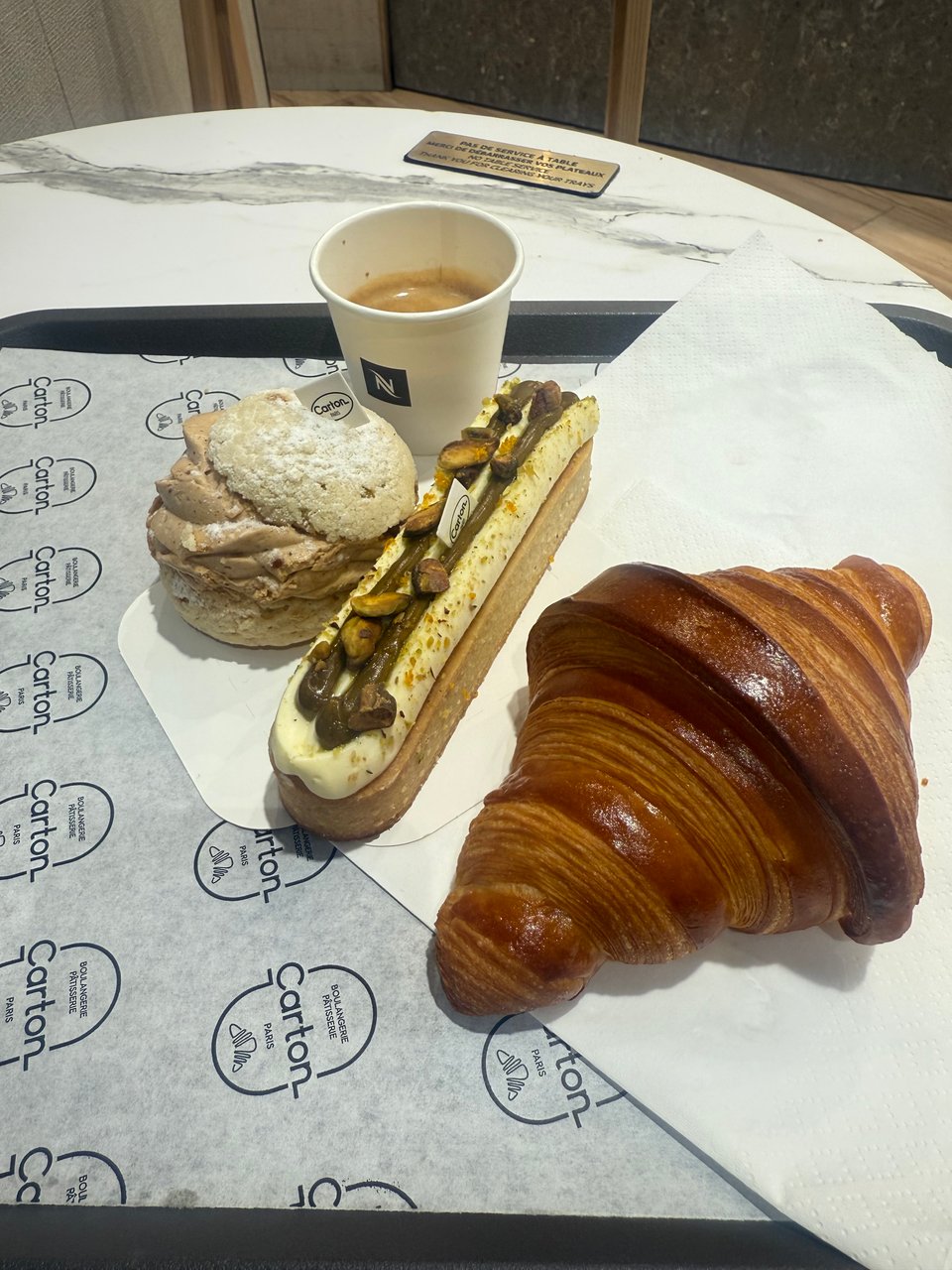 A photo of an espresso in a paper cup, a croissant, a Paris Brest, and a pistachio tarte.