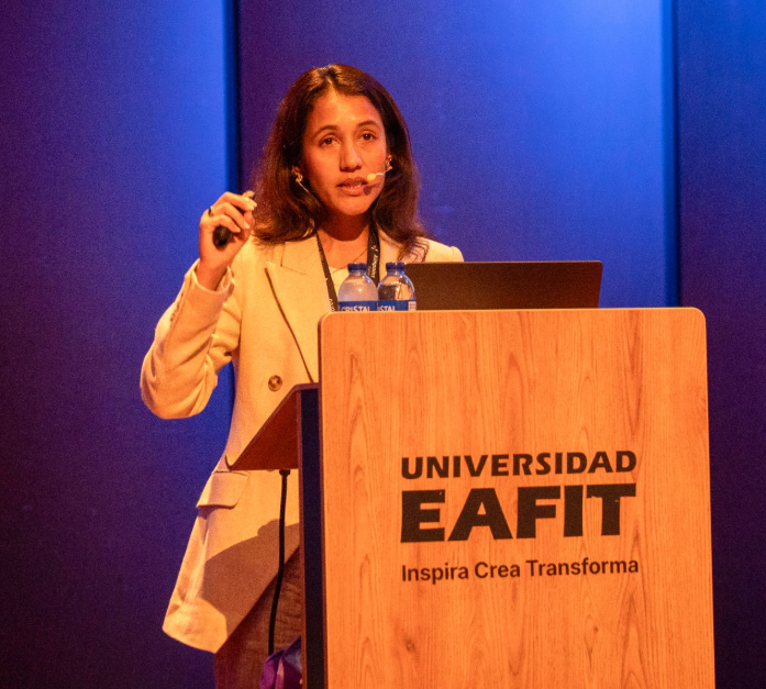 Photo of Malvika Sharan, an Indian women with long dark hair and a light coloured suit on, standing behind a lecturn giving a speech.