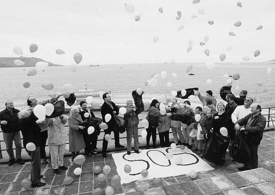 A group of people of all ages, standing on the roof top terrace and releasing balloons next to a handwritten sign on a sheet that spells SOS.