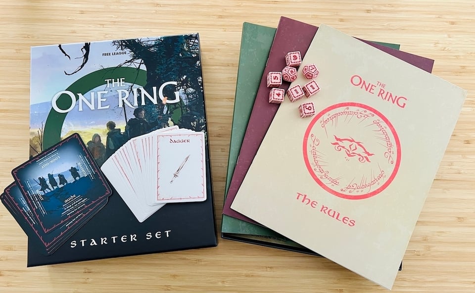 The box, dice, cards, and books of The One Ring Starter set spread on a wooden table.