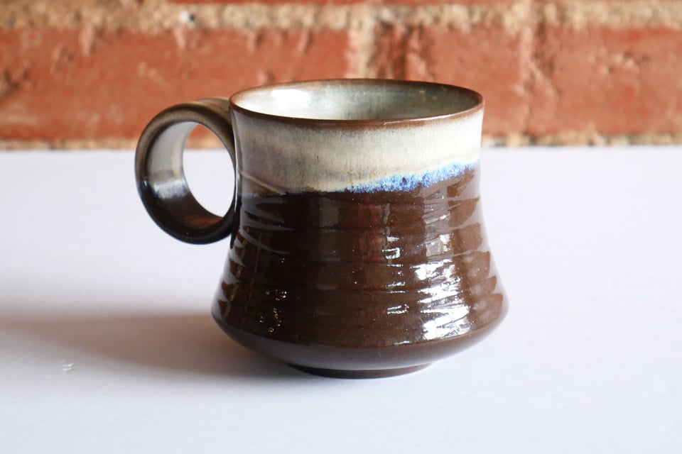 An inset foot and rounded base leads to curvy concave walls with textured grooves running around the exterior. The inside and outer rim is dipped at an angle in white glaze, which breaks brown over the lip and texture – fading to blues at the edges where it meats a clear glaze outside. The handle is a circle with room for a couple fingers, embedded into one side. It has a thin profile, but a wide surface that is comfortable on the hand.