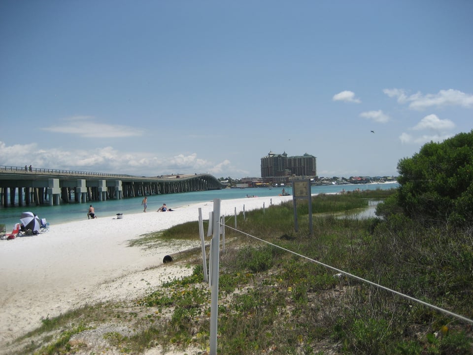 A white sand beach. A highway bridge crosses the inlet it faces, with a large building on the far side.