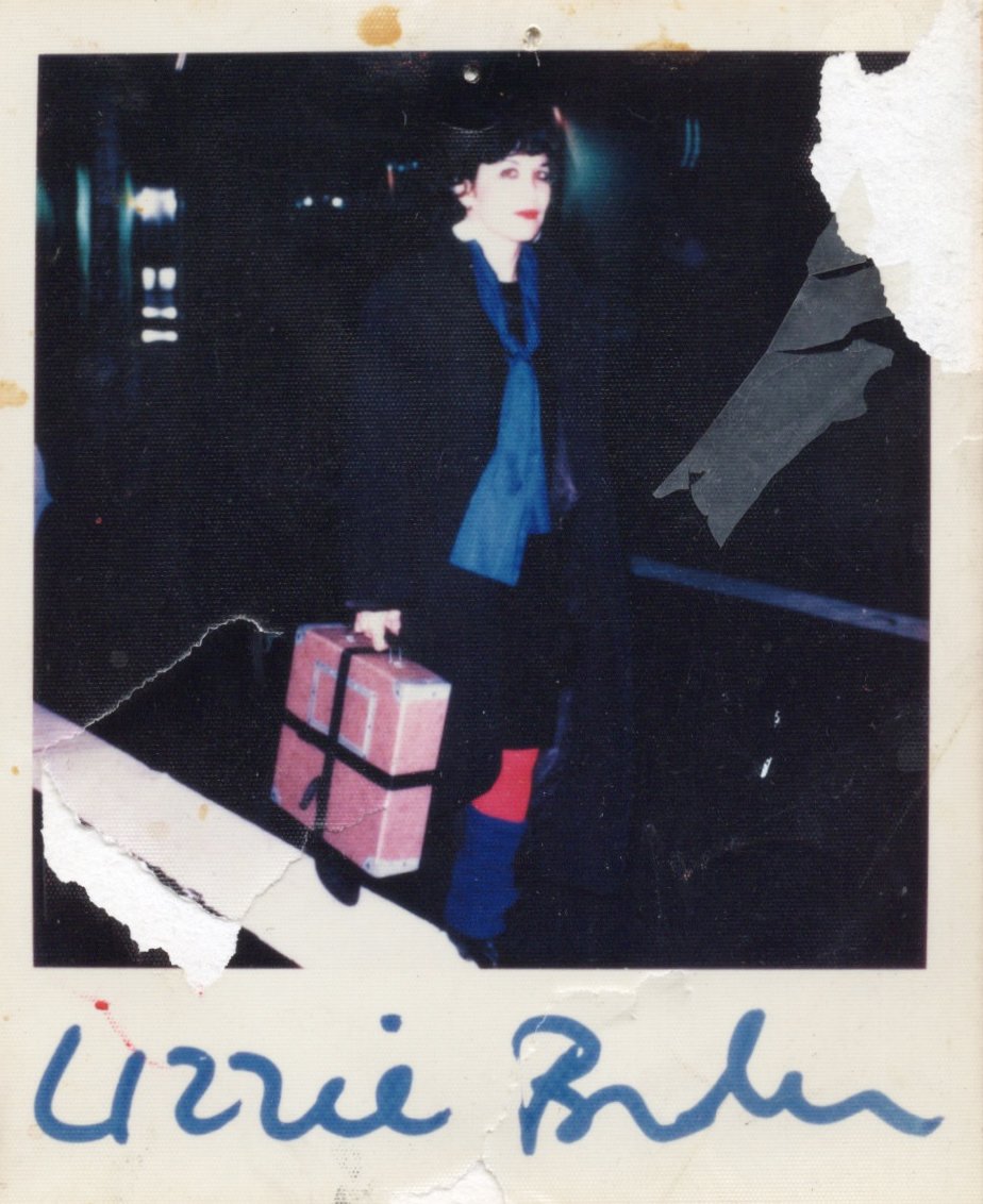 A worn Polaroid photograph showing a woman with short dark hair and red lipstick standing at night, holding a pink suitcase tied with black straps. She wears dark clothing with a blue scarf and red tights, and the photo’s edges are torn and stained, with a blue handwritten signature across the bottom that reads "Lizzie Borden"