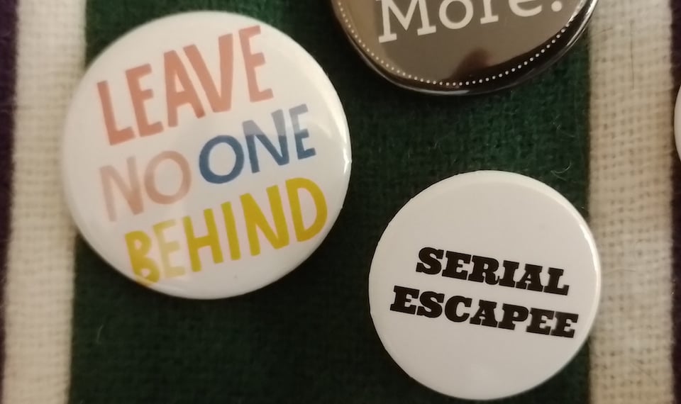 Two white button badges pinned to a striped scarf. The slightly larger badge on the left says Leave No One Behind in colourful capital letters. The smaller one on the right says Serial Escapee in black serif capitals.
