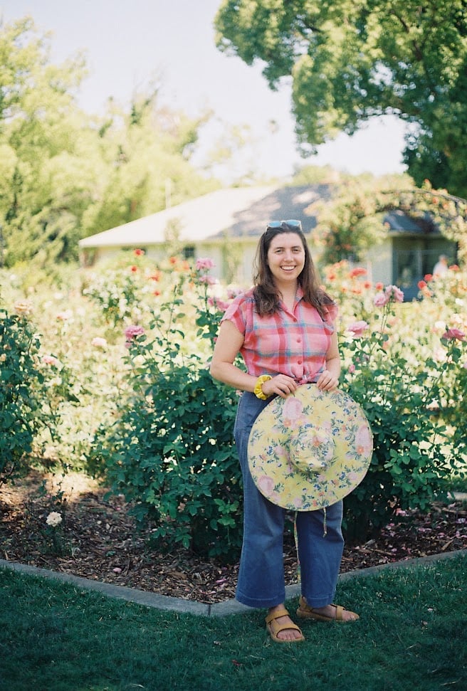 a white woman smiles, standing in front of a rose garden holding a sun hat
