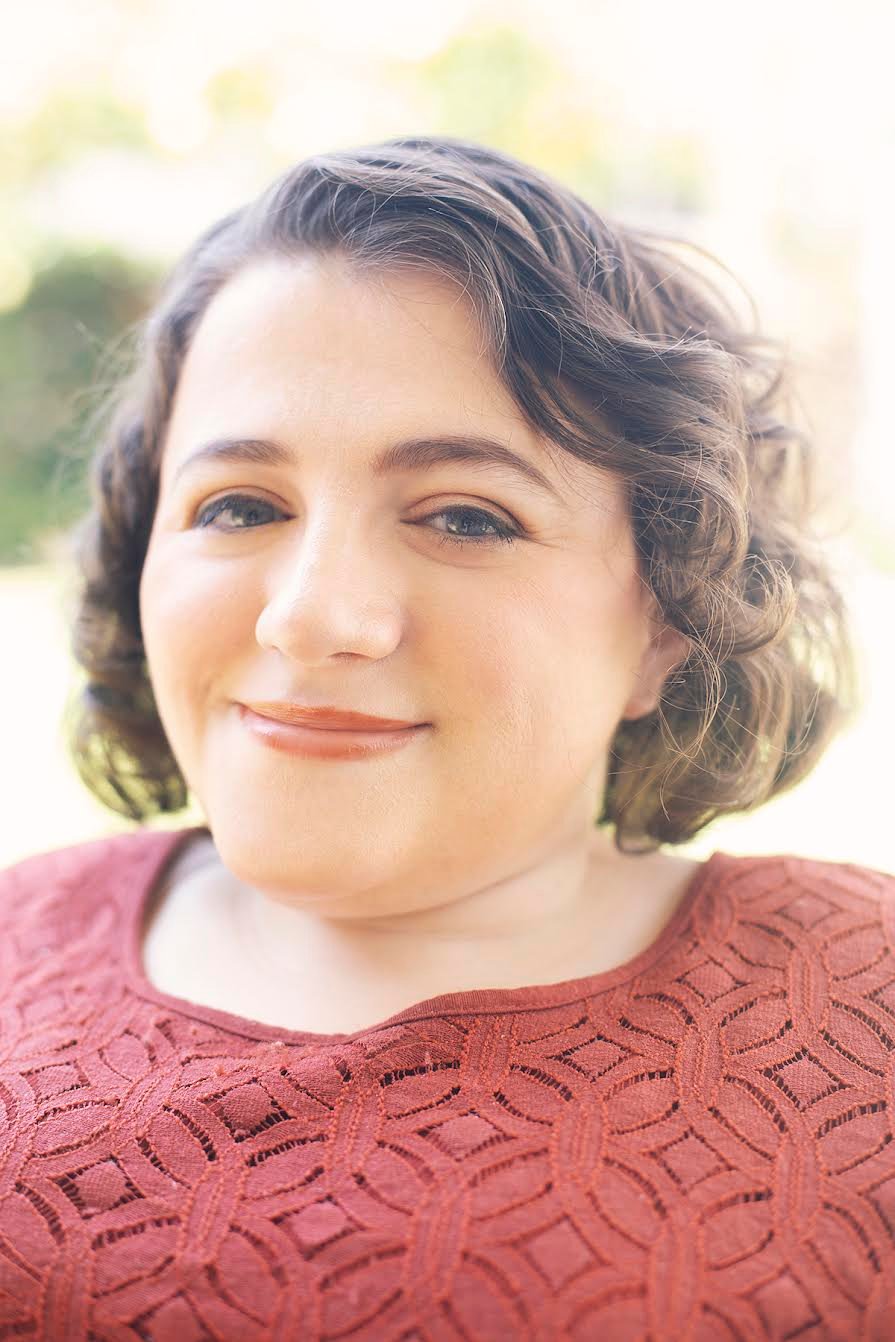 Photo of Kristen, a light-skinned woman with shoulder-length, wavy brown hair. She wears a brick red knit top and lipstick in the same color family.