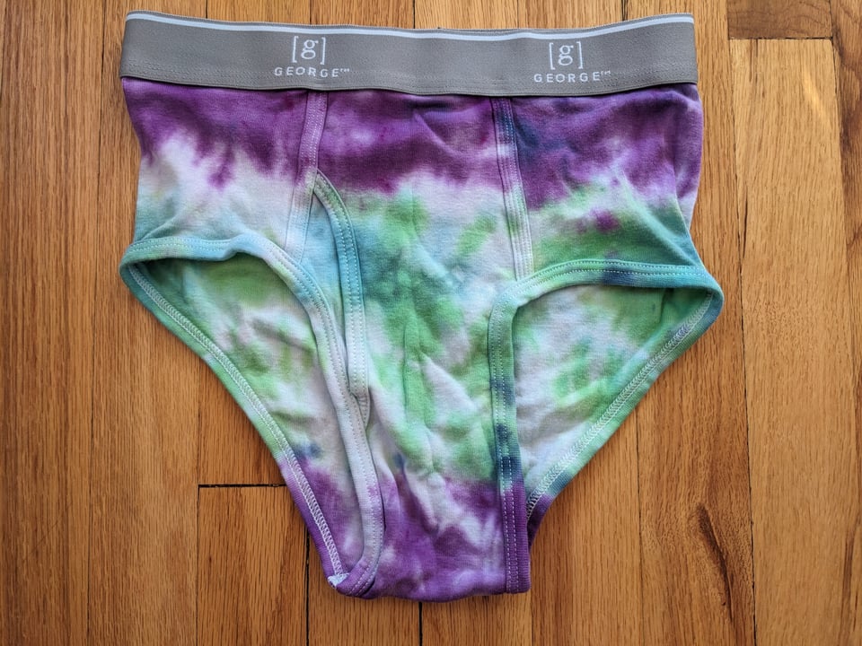 A pair of tie-dyed briefs, purple and green in wide stripes with a gray waistband, against a hardwood floor.