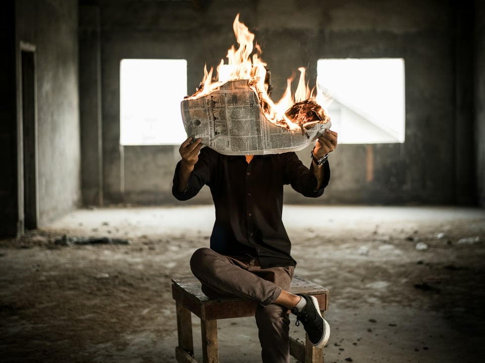 An image of a person, appearing to read a newspaper that is on fire. They are sitting in an empty room.