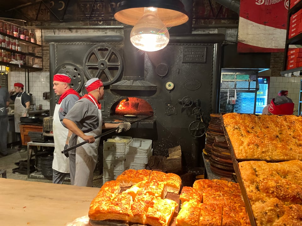Two cooks work in front of a large pizza oven while stacks of focaccia are shown in the foreground.