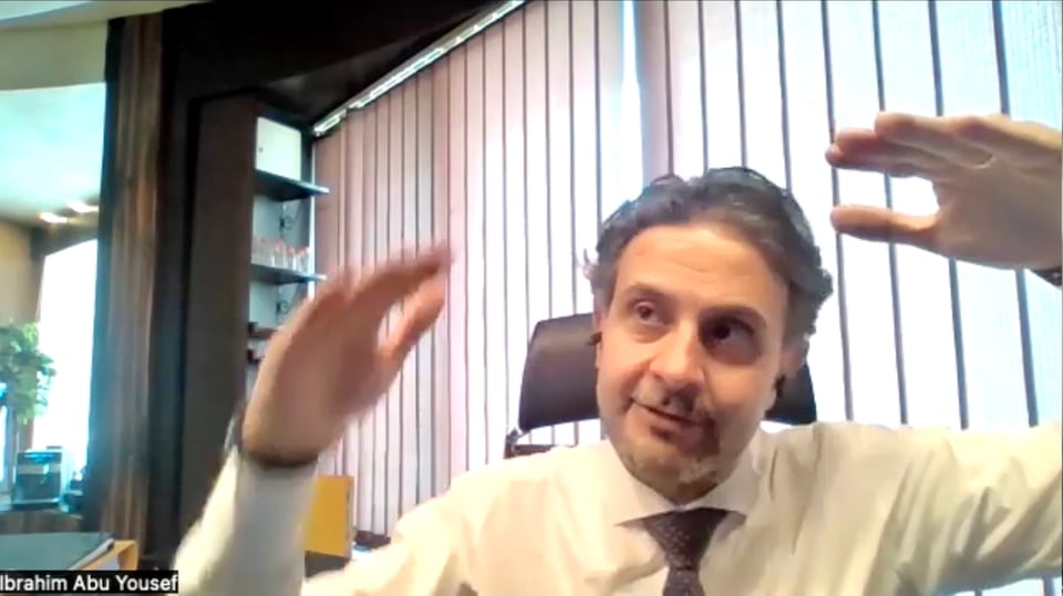 A man in shirt and tie sitting before a window with blinds closed, one of his raised hands just slightly lower than the other.