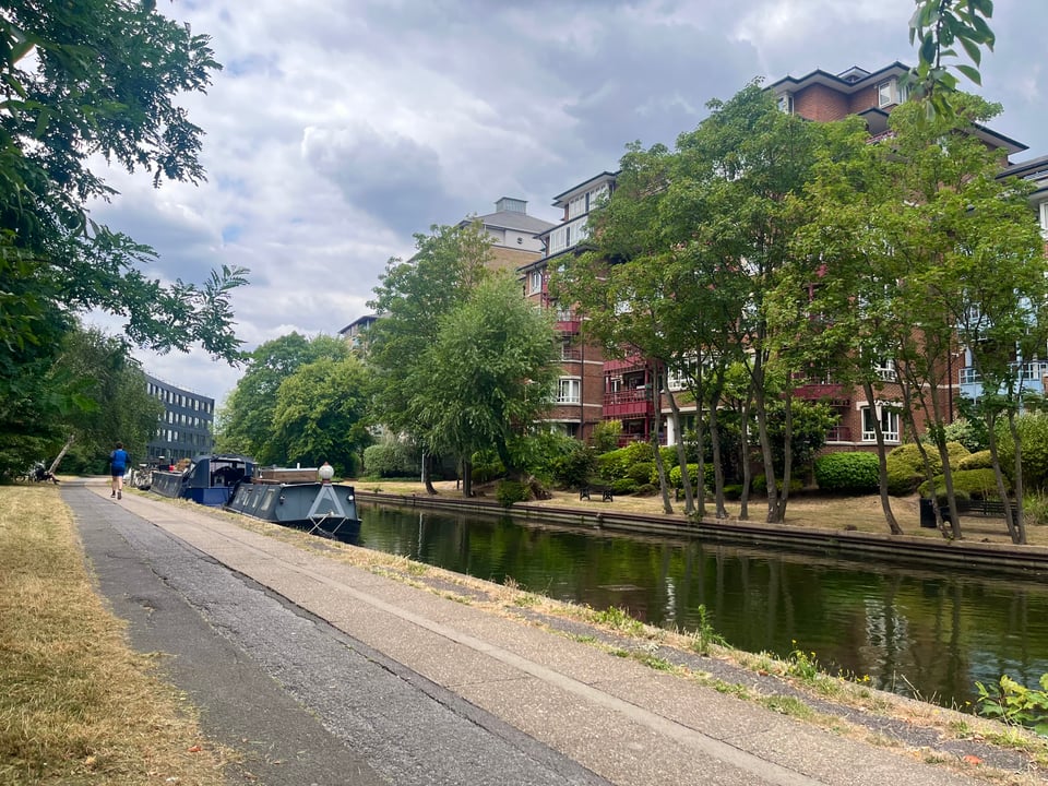 A serene canal in west London, with a few boats moored and trees on the other side. The sky is cloudy, which made the day pleasantly warm but not too sunny.
