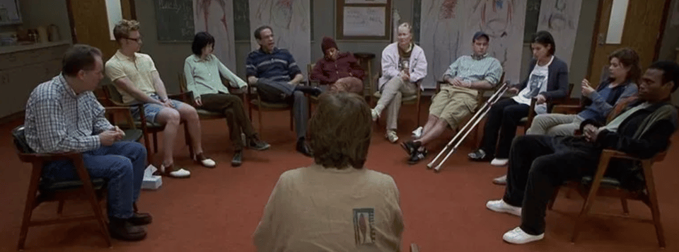 A still from 28 Days. 10 unhappy people sit in a semi-circle around a counsellor.