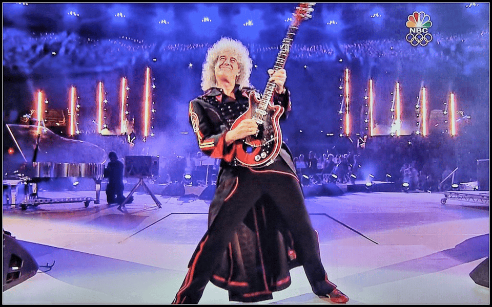 A photograph of the guitarist, Brian May on stage, playing his guitar. He is standing with his legs spread apart, and is leaning slightly back while holding his guitar upright, with a broad smile on his face as he leans his head backwards. In the far background of the stage are bright white lights, and someone playing a grand piano with a metal body and a translucent top.
