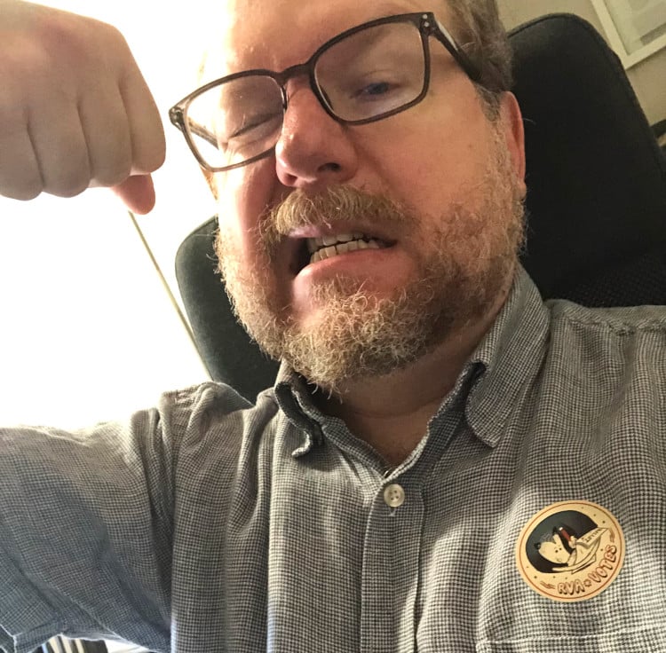 a white man with glasses and a gray beard, making a dorky expression. He wears a sticker with an opossum filling out a ballot on it, which he got for voting