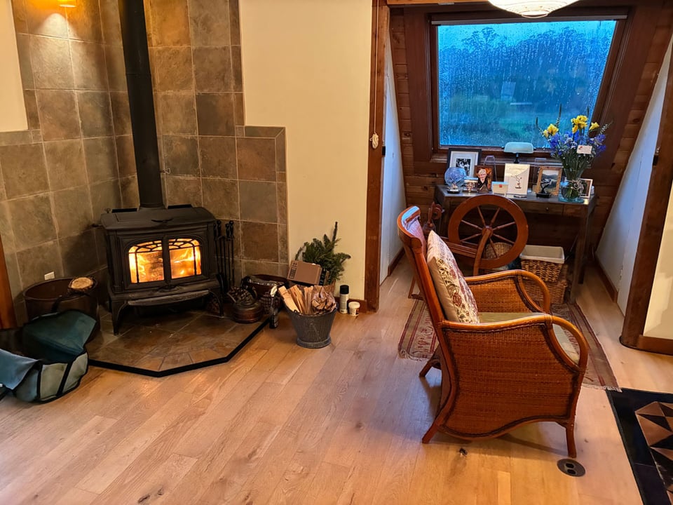 A cozy living room shows a fireplace with a fire lit. The window shows rain streaking down outside.
