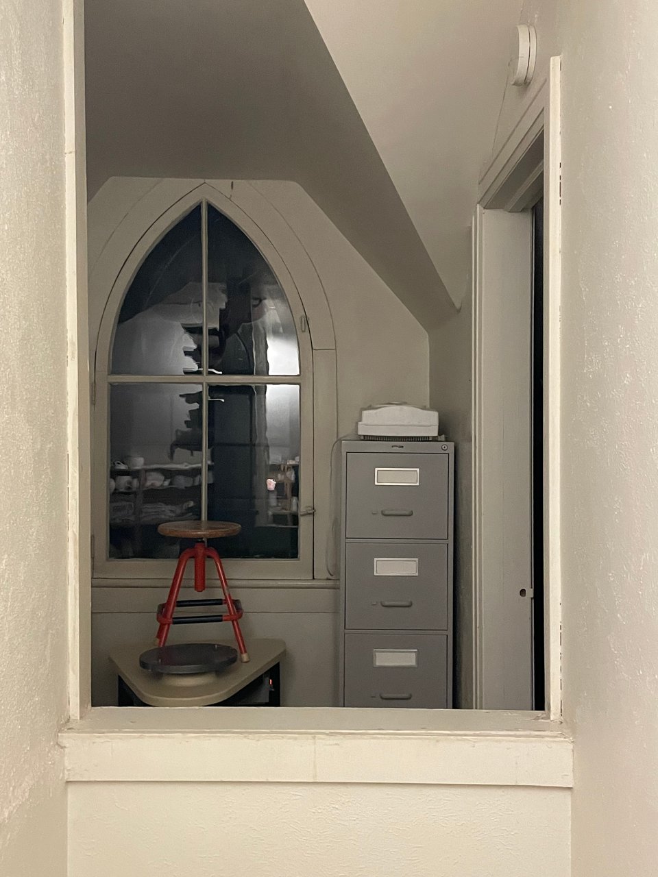 A wooden stool with red legs stands atop a pottery wheel table, on front of a darkened arched window. It is an old window has four panels, with a cross in the middle, forming an point at the top.Beside it sits a sturdy grey filing cabinet. White plaster walls frame the scene.