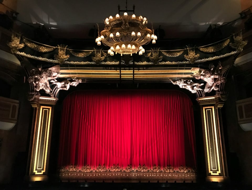 A long shot of a stage, with red curtains.  Photo by Gwen King on Unsplash.