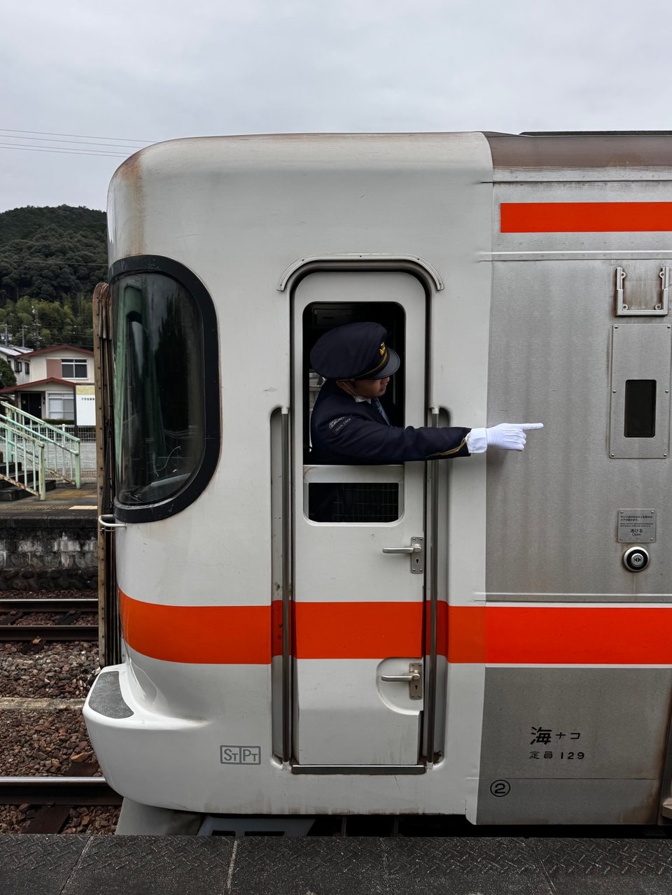 Train conductor leaning from the doorway of a silver and orange Japanese train, pointing along the platform.