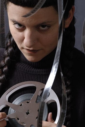 Wendy glares at the viewer, holding a film reel with film all over. Her hair is in braids and she wears a black turtleneck.