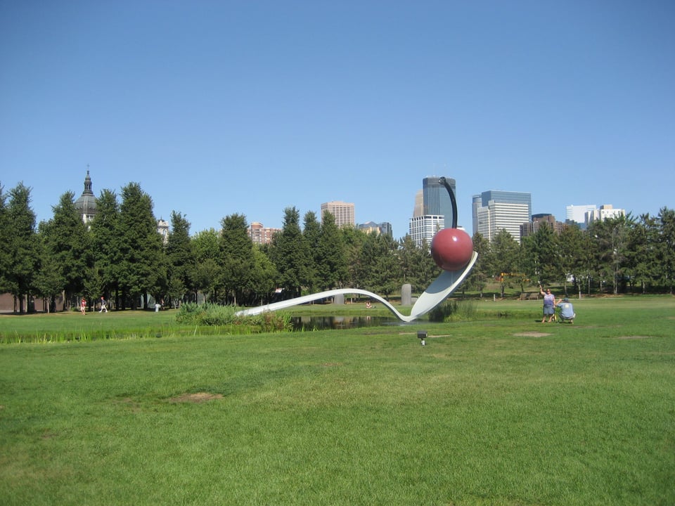 A sculpture of an extremely large spoon with a cherry on the tip. It's in a grassy park surrounded by trees and tall buildings.