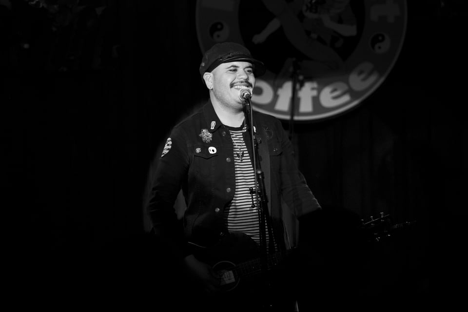 a man with a guitar, Marc-Alan Prince in front of a sign that reads Kick Butt Coffee in black and white live music photo