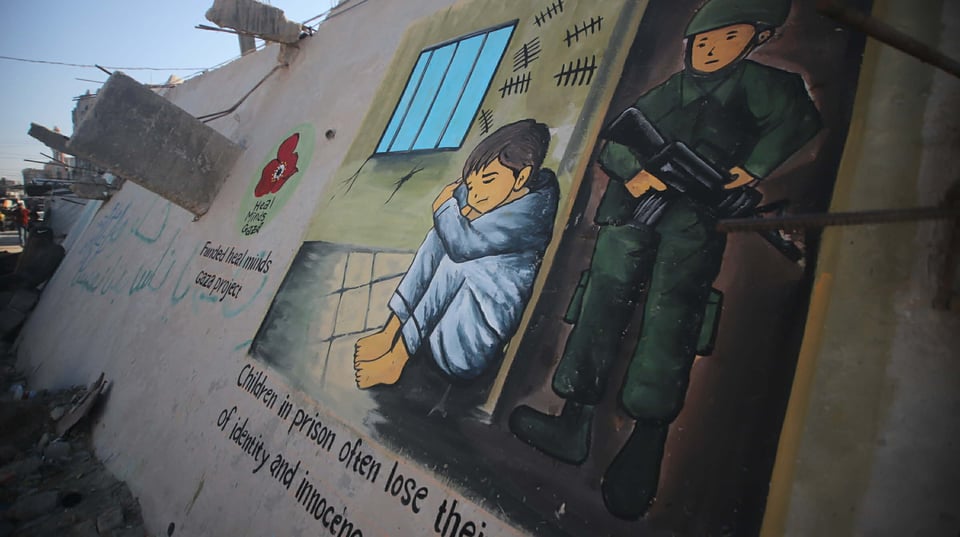 A perspective photo showing a painted mural on a wall in Gaza, illustrating a child curled up on a prison cell floor next to a barred window and tally marks drawn on the wall. In an open doorway behind the child is an imposing soldier with a machine gun. Below the mural is some cropped text: "Children in prison often lose their sense of identity and innocence." To the side of the mural is the Heal Minds Gaza logo with the text "funded by Heal Minds Gaza project."