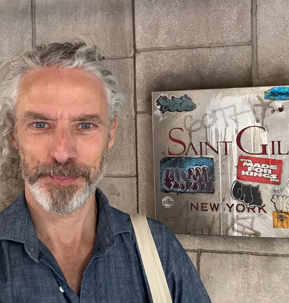 Selfie of silver-haired white man in dark denim shirt standing next to a graffiti-covered sign that reads SAINT GIL