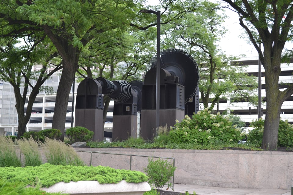 A black sculpture of pulley wheels on gray concrete stands, surrounded by trees.