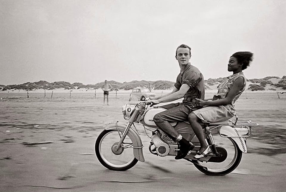 A black and white photograph of a couple on a motorcycle riding over a vast beach. The boy is in front and looking toward the camera. The girl is in the back, holding on to his waist, her short hair blowing behind her.